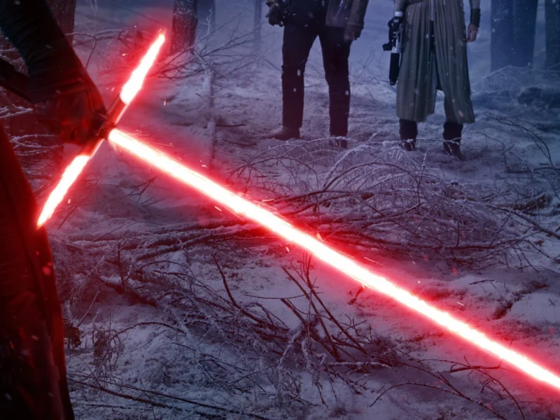 Un laser sabre crossguard rouge brillant se trouve sur un sol couvert de givre dans une forêt enneigée, avec deux personnages sombres debout en arrière-plan.