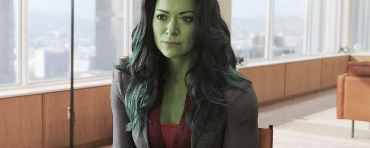 Une femme à la peau verte, assise à une table dans un bureau moderne avec un paysage urbain visible à travers une grande fenêtre, portant un blazer gris et un chemisier rouge, ressemblant au personnage de Marvel She-Hulk
