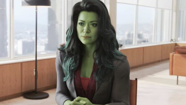 Une femme à la peau verte, assise à une table dans un bureau moderne avec un paysage urbain visible à travers une grande fenêtre, portant un blazer gris et un chemisier rouge, ressemblant au personnage de Marvel She-Hulk