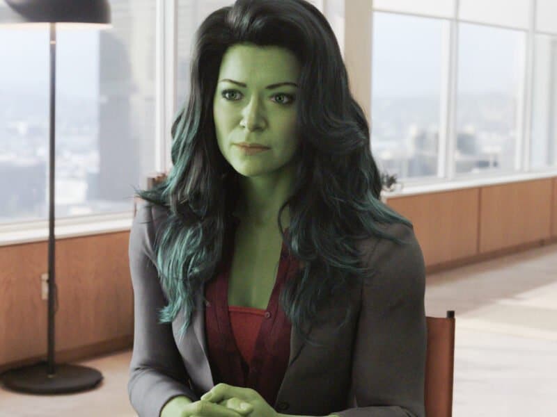 Une femme à la peau verte, assise à une table dans un bureau moderne avec un paysage urbain visible à travers une grande fenêtre, portant un blazer gris et un chemisier rouge, ressemblant au personnage de Marvel She-Hulk