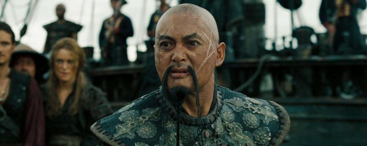 Gros plan d'un homme chauve au regard intense, portant une armure aux motifs élaborés, debout sur le pont d'un navire entouré d'autres personnages Disney.