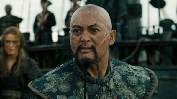 Gros plan d'un homme chauve au regard intense, portant une armure aux motifs élaborés, debout sur le pont d'un navire entouré d'autres personnages Disney.