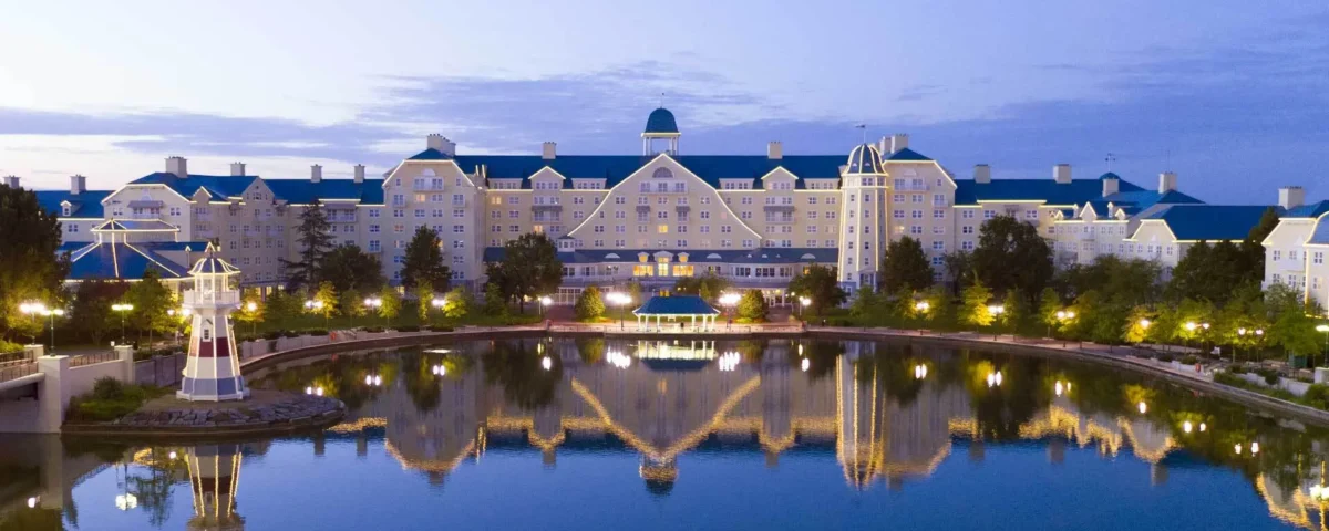 Une vue panoramique d'un grand hôtel de style complexe Disneyland Paris Newport Bay Club avec des toits bleus, qui se reflète magnifiquement dans un plan d'eau serein au crépuscule. Une petite structure de phare est également visible.