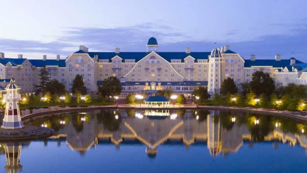 Une vue panoramique d'un grand hôtel de style complexe Disneyland Paris Newport Bay Club avec des toits bleus, qui se reflète magnifiquement dans un plan d'eau serein au crépuscule. Une petite structure de phare est également visible.