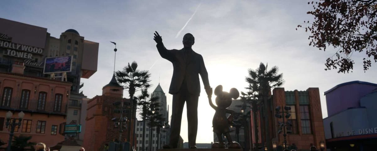 Statue de Walt Disney serrant les mains de Mickey Mouse, se découpant sur un ciel lumineux, avec des attractions de parc d'attractions en arrière-plan.