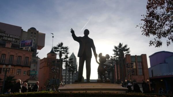 Statue de Walt Disney serrant les mains de Mickey Mouse, se découpant sur un ciel lumineux, avec des attractions de parc d'attractions en arrière-plan.