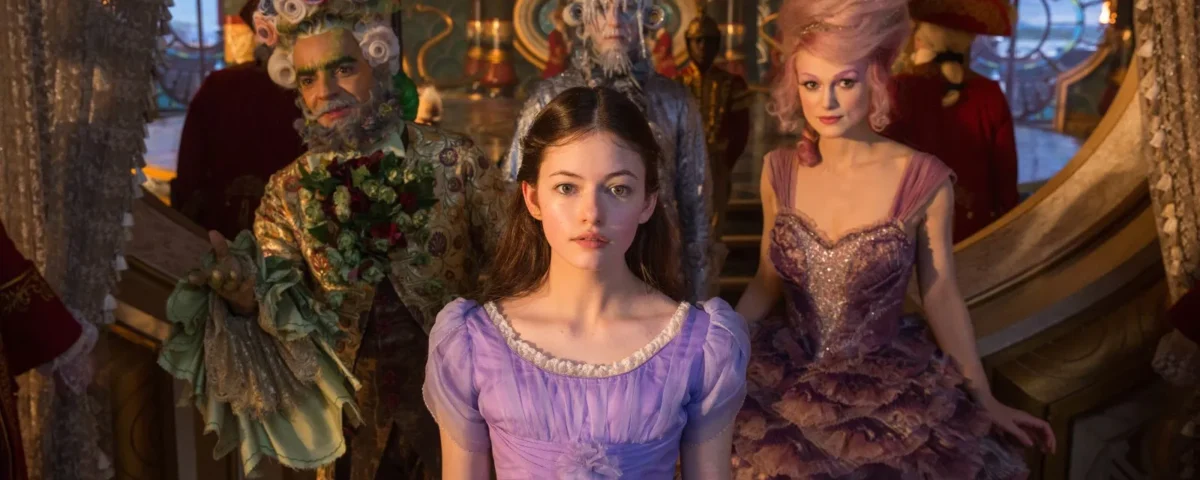 Une jeune femme vêtue d'une robe lavande se tient au centre, entourée de trois personnages fantaisistes vêtus de costumes de casse-noisette élaborés et colorés, dans un environnement fantastique et richement décoré.