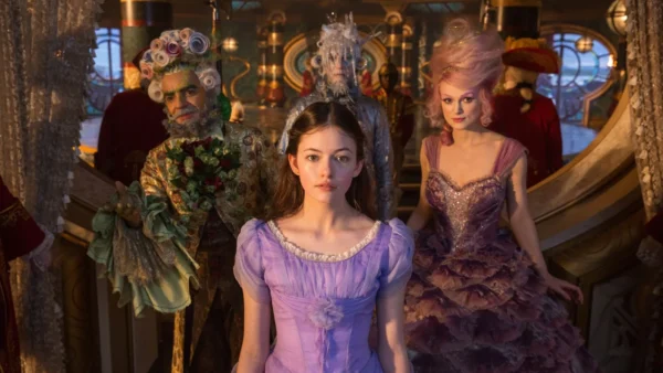Une jeune femme vêtue d'une robe lavande se tient au centre, entourée de trois personnages fantaisistes vêtus de costumes de casse-noisette élaborés et colorés, dans un environnement fantastique et richement décoré.