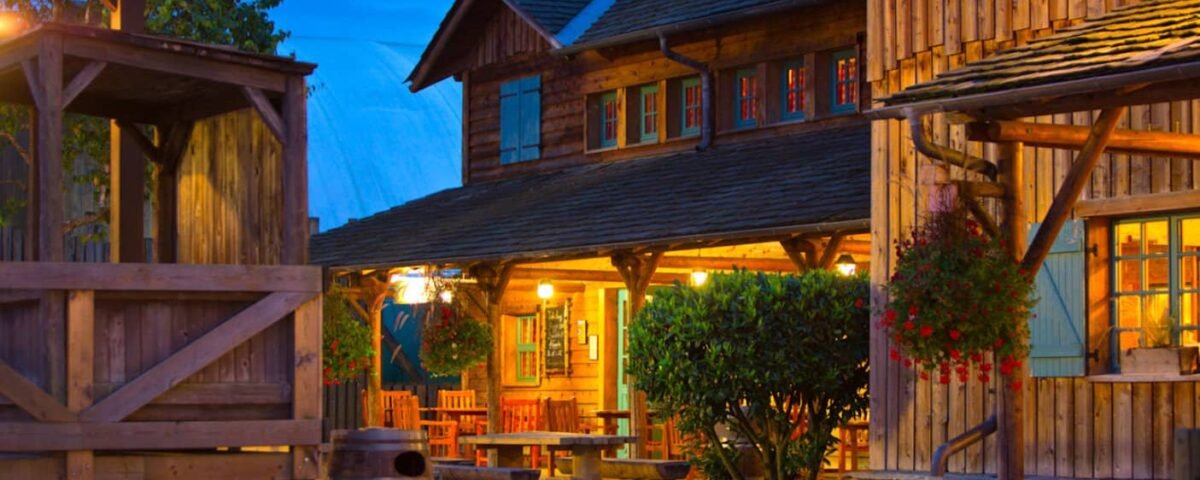Une cabane en bois pittoresque et une structure adjacente au Davy Crockett Ranch, toutes deux chaleureusement éclairées, entourées de verdure luxuriante et de fleurs au crépuscule dans un village rustique à l'ancienne près de Disneyland Paris.