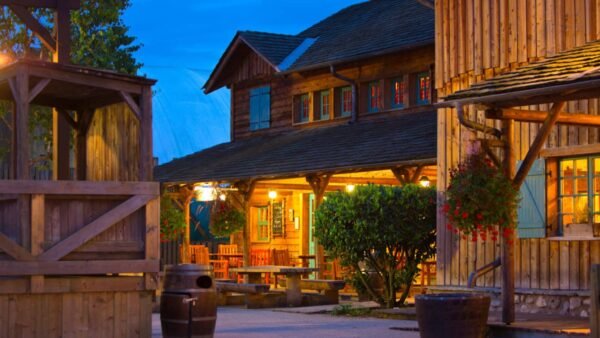 Une cabane en bois pittoresque et une structure adjacente au Davy Crockett Ranch, toutes deux chaleureusement éclairées, entourées de verdure luxuriante et de fleurs au crépuscule dans un village rustique à l'ancienne près de Disneyland Paris.