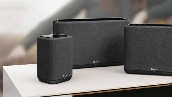 Trois enceintes Wi-Fi Denon modernes recouvertes de tissu noir, exposées sur une table en bois dans une pièce élégamment décorée.
