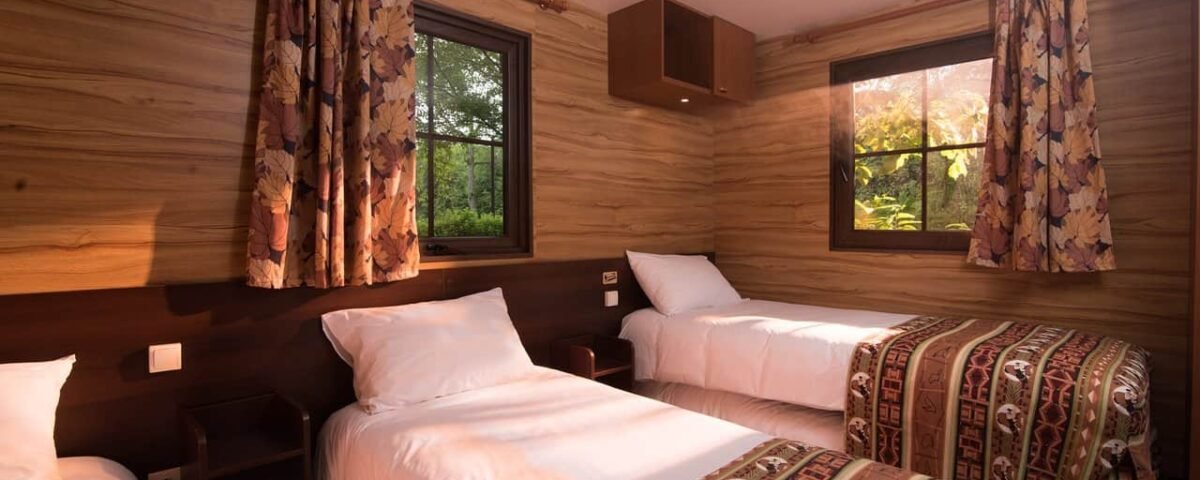 Une cabine confortable en bois au Davy Crockett Ranch avec deux lits simples recouverts de couvertures à motifs, une table de chevet, des rideaux aux fenêtres et une vue sur la verdure à l'extérieur.