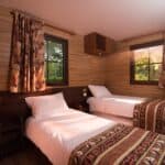 Une cabine confortable en bois au Davy Crockett Ranch avec deux lits simples recouverts de couvertures à motifs, une table de chevet, des rideaux aux fenêtres et une vue sur la verdure à l'extérieur.