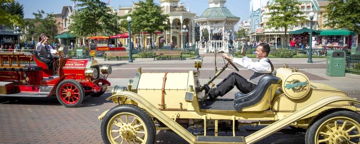 Une scène animée à Disneyland Paris montrant un homme conduisant une voiture jaune à l'ancienne, saluant joyeusement tandis qu'un véhicule vintage rouge passe dans la direction opposée par une journée ensoleillée.