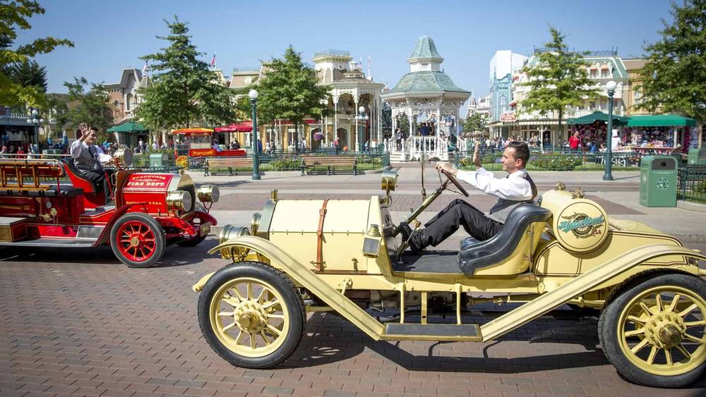 Une scène animée à Disneyland Paris montrant un homme conduisant une voiture jaune à l'ancienne, saluant joyeusement tandis qu'un véhicule vintage rouge passe dans la direction opposée par une journée ensoleillée.