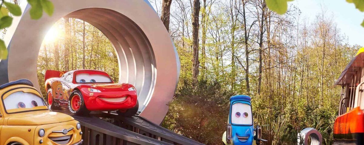 Des voitures animées du film "Cars" entrant dans un grand portail en forme d'aimant sur une route forestière ensoleillée à Disneyland Paris, avec le soleil se couchant en arrière-plan.