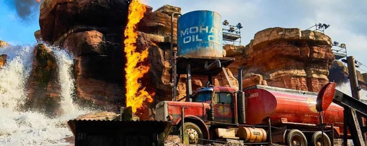 Une scène dynamique lors d'un spectacle de cascades à Disneyland mettant en vedette une grande cascade, un terrain accidenté et un camion-citerne rouge garé près d'un château d'eau "Mohawk Oil" avec des flammes éclatant à côté.