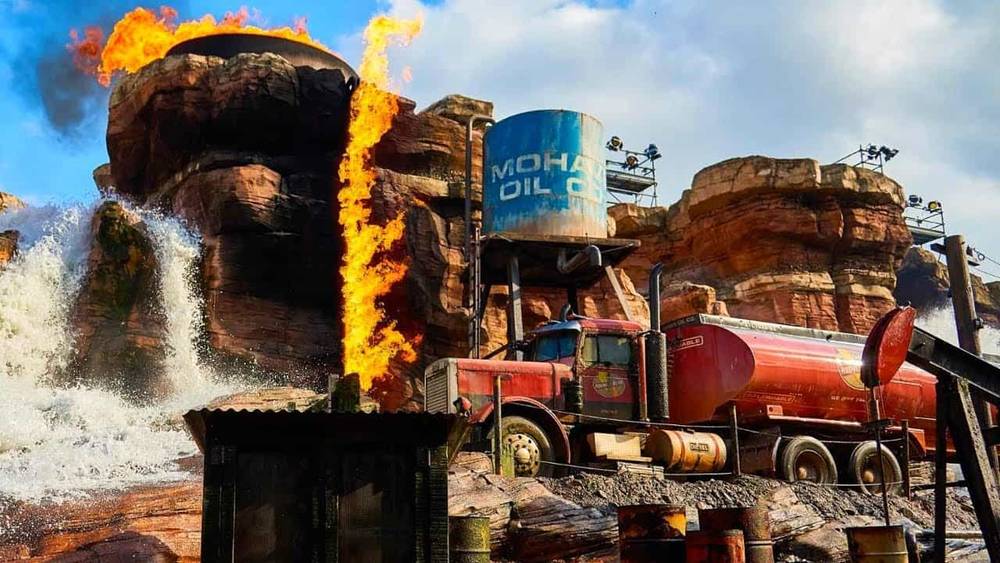 Une scène dynamique lors d'un spectacle de cascades à Disneyland mettant en vedette une grande cascade, un terrain accidenté et un camion-citerne rouge garé près d'un château d'eau "Mohawk Oil" avec des flammes éclatant à côté.