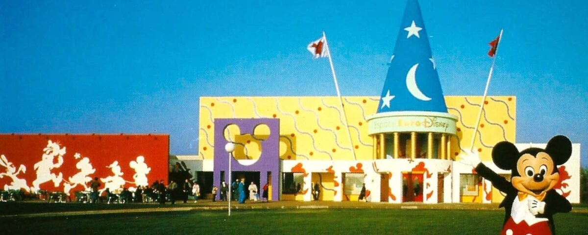 Un joyeux personnage de Mickey Mouse se tient devant l'entrée colorée de l'Espace Euro Disney, avec des motifs et des drapeaux fantaisistes, sous un ciel bleu clair.