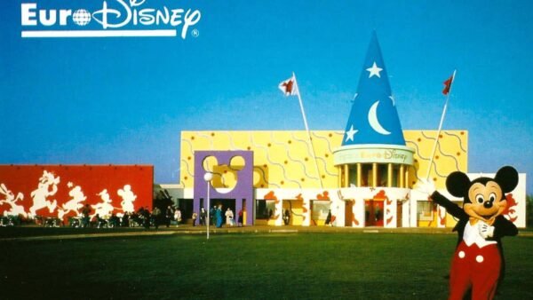 Un joyeux personnage de Mickey Mouse se tient devant l'entrée colorée de l'Espace Euro Disney, avec des motifs et des drapeaux fantaisistes, sous un ciel bleu clair.