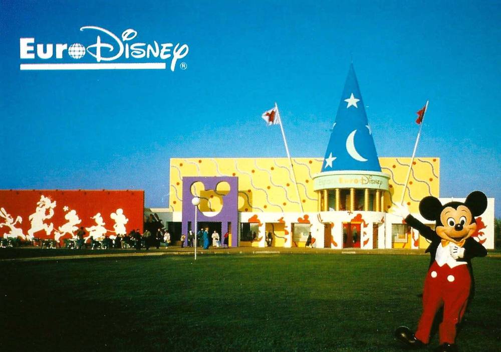 Un joyeux personnage de Mickey Mouse se tient devant l'entrée colorée de l'Espace Euro Disney, avec des motifs et des drapeaux fantaisistes, sous un ciel bleu clair.