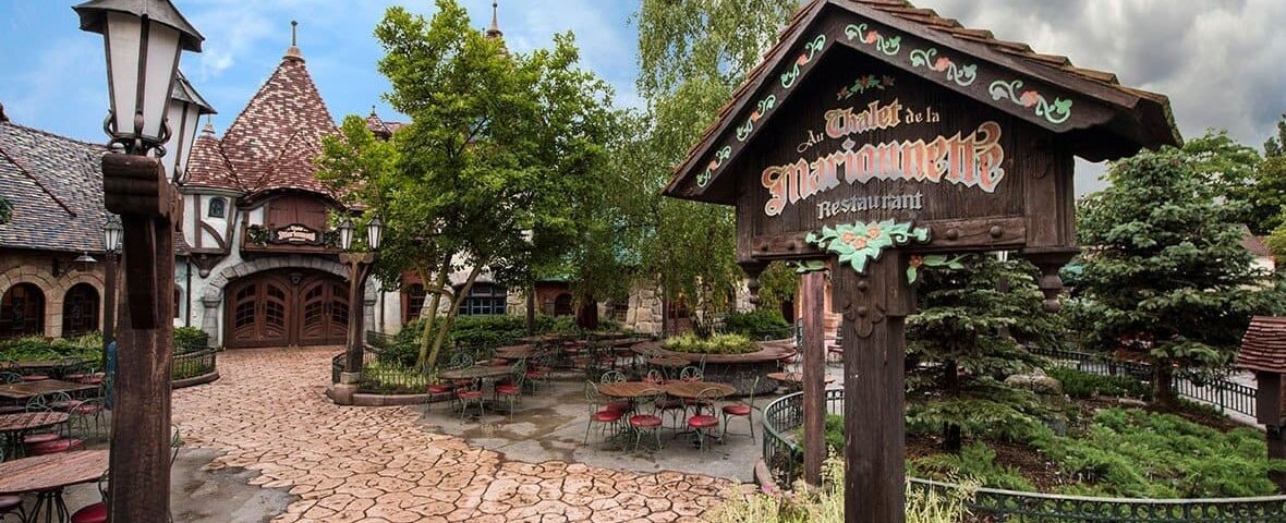 Une cour de restaurant pittoresque sur le thème médiéval à Disneyland avec des allées pavées, des tables rustiques en bois et une pancarte décorative indiquant "Auberge de Cendrillon". Des arbres verdoyants et