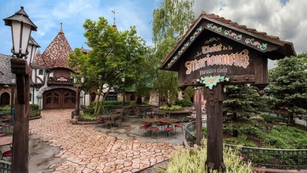 Une cour de restaurant pittoresque sur le thème médiéval à Disneyland avec des allées pavées, des tables rustiques en bois et une pancarte décorative indiquant "Auberge de Cendrillon". Des arbres verdoyants et