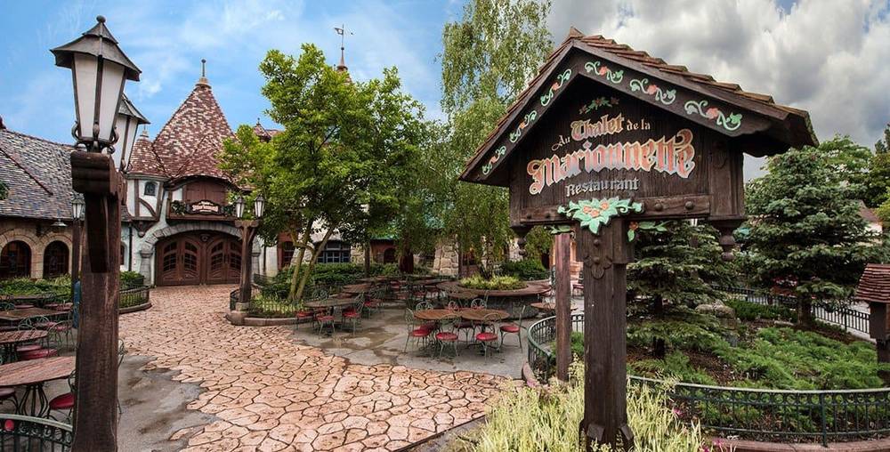 Une cour de restaurant pittoresque sur le thème médiéval à Disneyland avec des allées pavées, des tables rustiques en bois et une pancarte décorative indiquant "Auberge de Cendrillon". Des arbres verdoyants et
