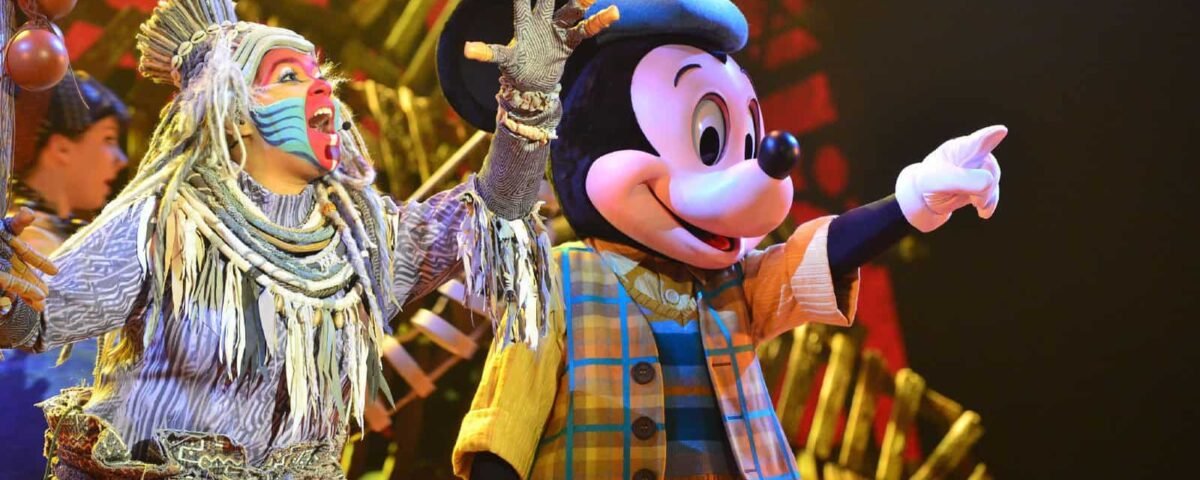 Mickey Mouse, vêtu d'un gilet à carreaux et de gants, interagit joyeusement avec un artiste vêtu d'un costume tribal coloré sur une scène aux couleurs vives de Disneyland.