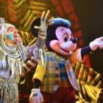 Mickey Mouse, vêtu d'un gilet à carreaux et de gants, interagit joyeusement avec un artiste vêtu d'un costume tribal coloré sur une scène aux couleurs vives de Disneyland.