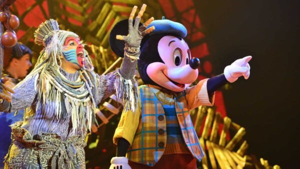 Mickey Mouse, vêtu d'un gilet à carreaux et de gants, interagit joyeusement avec un artiste vêtu d'un costume tribal coloré sur une scène aux couleurs vives de Disneyland.