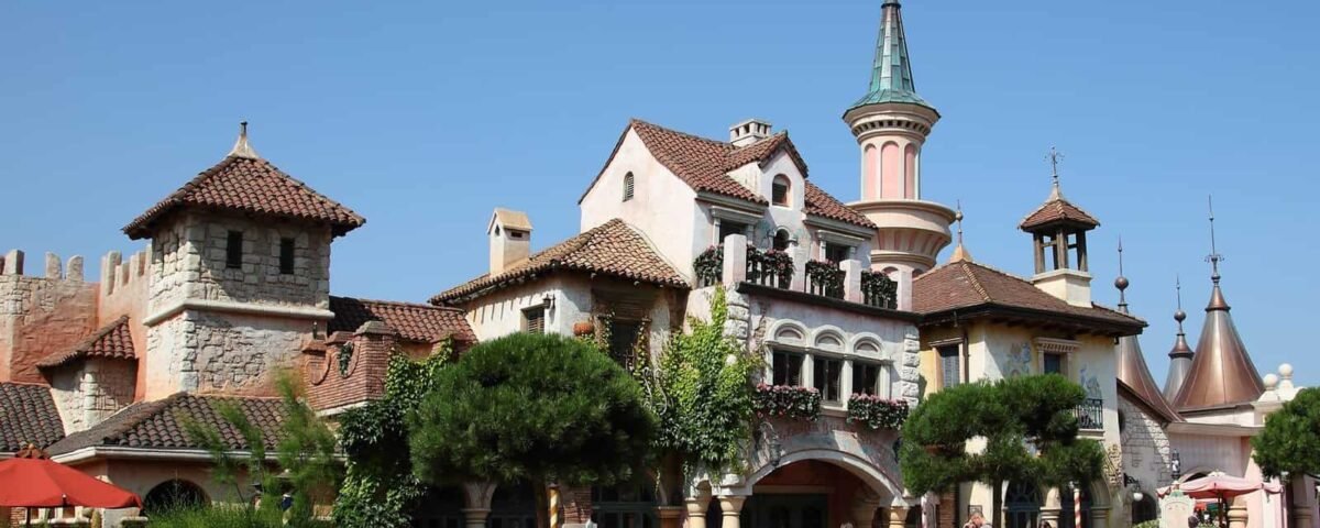 Vue ensoleillée d'un charmant village de style européen avec des bâtiments roses et beiges ornés de verdure, avec des tourelles pittoresques et des toits de tuiles rouges. Les visiteurs déambulent sous un ciel bleu clair près de la Bella Notte