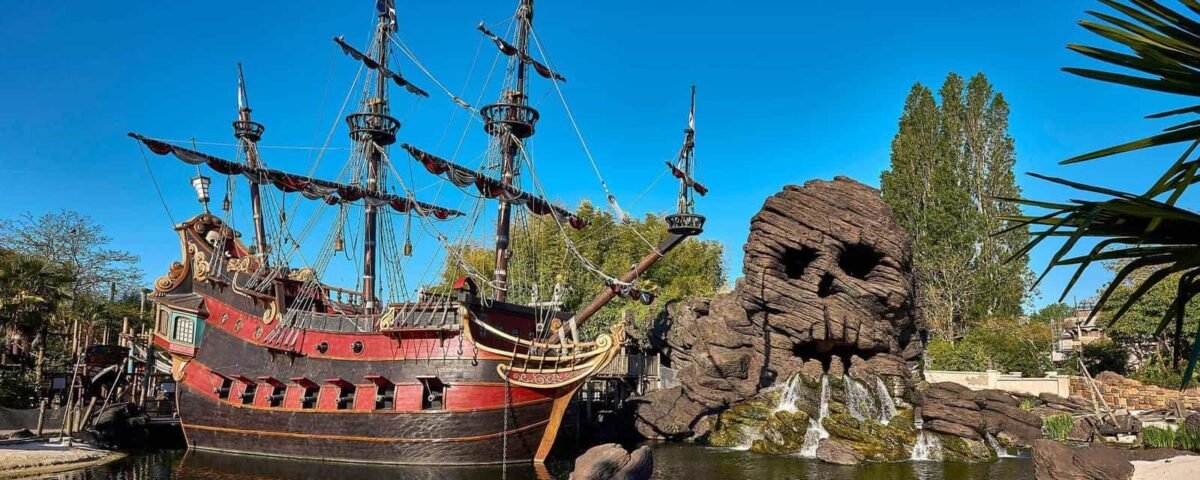Un grand bateau pirate orné rappelant les aventures de Peter Pan, avec plusieurs mâts ancrés dans un petit lac à côté d'un éperon rocheux avec des cascades et de la verdure sous un ciel bleu clair