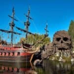 Un grand bateau pirate orné rappelant les aventures de Peter Pan, avec plusieurs mâts ancrés dans un petit lac à côté d'un éperon rocheux avec des cascades et de la verdure sous un ciel bleu clair