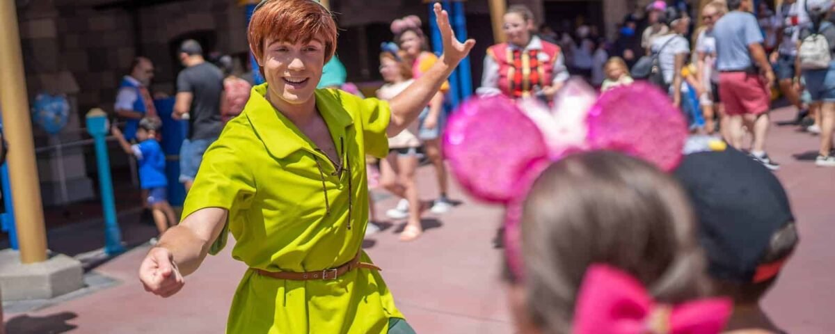 Un artiste habillé en Peter Pan accueille les visiteurs avec un grand sourire et un signe de la main à Disneyland, avec des gens en arrière-plan profitant de la journée ensoleillée.