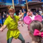 Un artiste habillé en Peter Pan accueille les visiteurs avec un grand sourire et un signe de la main à Disneyland, avec des gens en arrière-plan profitant de la journée ensoleillée.