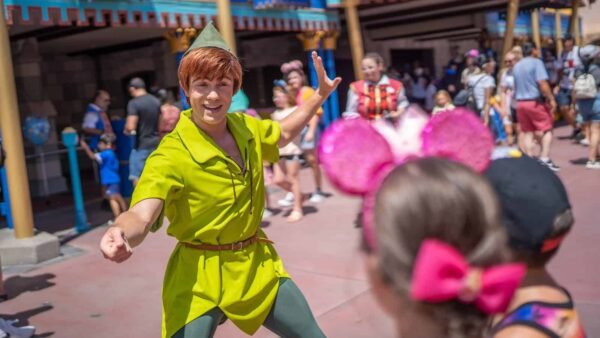 Un artiste habillé en Peter Pan accueille les visiteurs avec un grand sourire et un signe de la main à Disneyland, avec des gens en arrière-plan profitant de la journée ensoleillée.