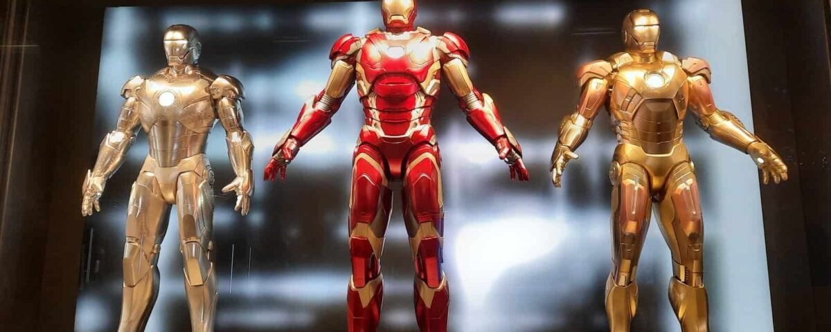 Trois combinaisons d'Iron Man exposées dans un hôtel : une argentée, une rouge et dorée au centre et une dorée, le tout placé contre un mur rétro-éclairé dans une pièce sombre.