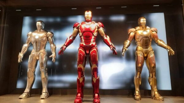 Trois combinaisons d'Iron Man exposées dans un hôtel : une argentée, une rouge et dorée au centre et une dorée, le tout placé contre un mur rétro-éclairé dans une pièce sombre.