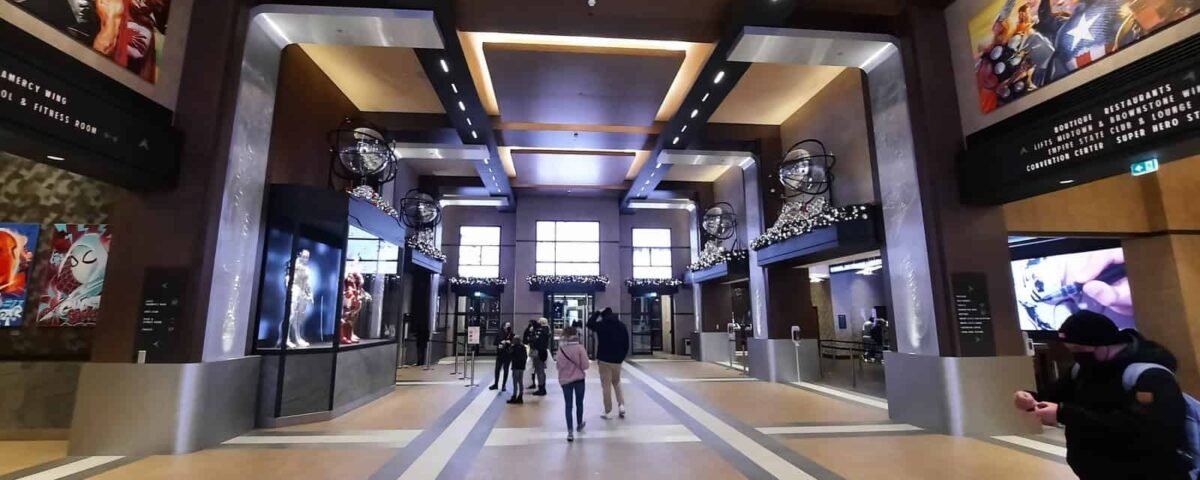 Hall de cinéma spacieux au design moderne avec des passants, des affiches de films sur les murs et des écrans numériques affichant les horaires des films. Un éclairage lumineux et directionnel et des détails architecturaux élégants rappelant Disneyland Paris.