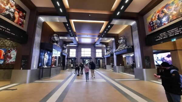 Hall de cinéma spacieux au design moderne avec des passants, des affiches de films sur les murs et des écrans numériques affichant les horaires des films. Un éclairage lumineux et directionnel et des détails architecturaux élégants rappelant Disneyland Paris.