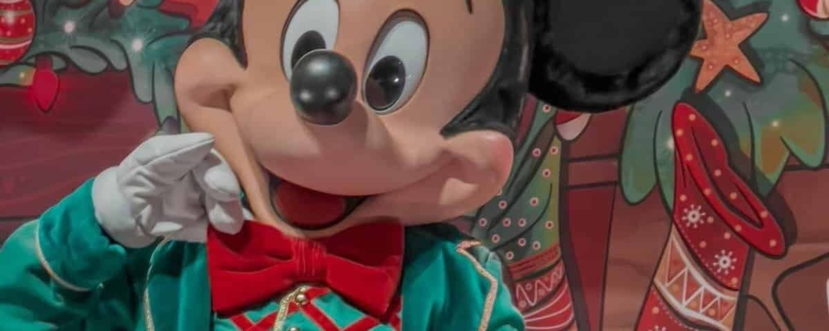 Mickey Mouse dans une tenue verte festive avec un nœud papillon rouge, tenant un petit cadeau, debout devant un décor coloré sur le thème de Noël avec des bas et des ornements.