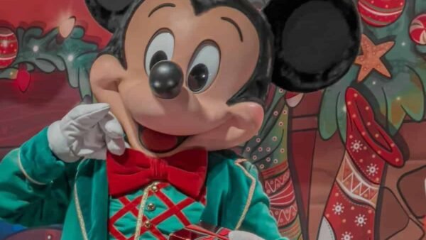 Mickey Mouse dans une tenue verte festive avec un nœud papillon rouge, tenant un petit cadeau, debout devant un décor coloré sur le thème de Noël avec des bas et des ornements.