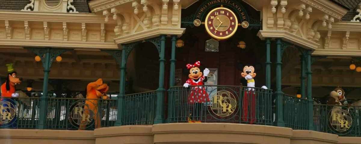 Minnie Mouse et Mickey Mouse se tiennent sur un balcon au-dessus de l'entrée de Disneyland, flanqués de Dingo et Pluto, alors qu'ils accueillent les visiteurs. La façade présente des détails ornés et un grand coul