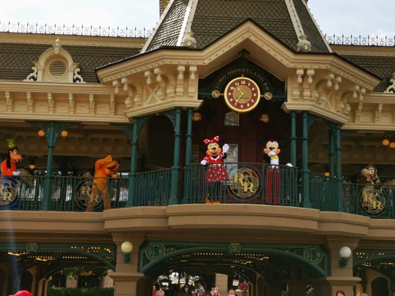 Minnie Mouse et Mickey Mouse se tiennent sur un balcon au-dessus de l'entrée de Disneyland, flanqués de Dingo et Pluto, alors qu'ils accueillent les visiteurs. La façade présente des détails ornés et un grand coul