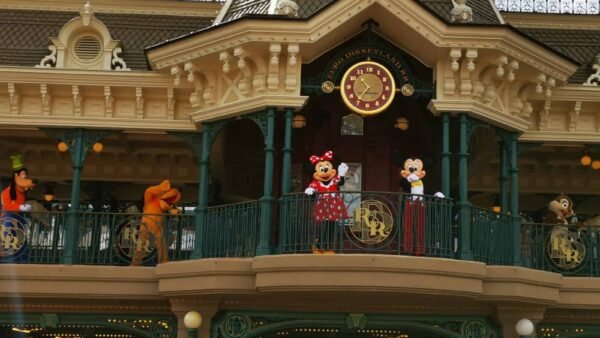 Minnie Mouse et Mickey Mouse se tiennent sur un balcon au-dessus de l'entrée de Disneyland, flanqués de Dingo et Pluto, alors qu'ils accueillent les visiteurs. La façade présente des détails ornés et un grand coul