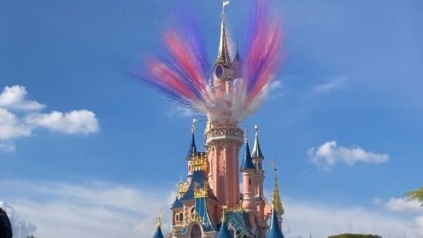 Un spectacle coloré de fumée ressemblant à un feu d'artifice éclate au-dessus de l'emblématique château de la Belle au bois dormant à Disneyland Paris sous un ciel bleu clair.