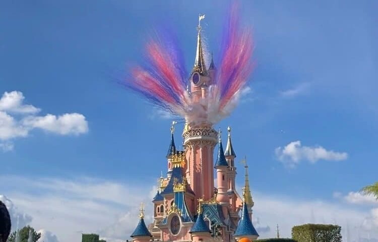 Un spectacle coloré de fumée ressemblant à un feu d'artifice éclate au-dessus de l'emblématique château de la Belle au bois dormant à Disneyland Paris sous un ciel bleu clair.