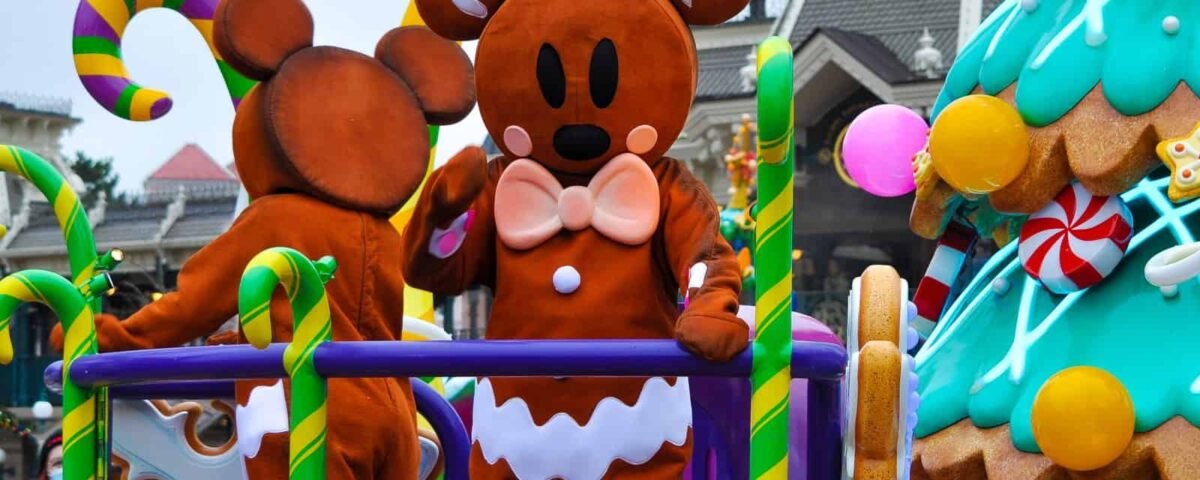 Une personne vêtue d'un costume de pain d'épice festif avec des accents de canne en bonbon salue un char coloré orné de bonbons et d'une maison en pain d'épice sur le thème de Noël.