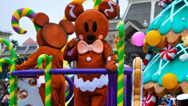 Une personne vêtue d'un costume de pain d'épice festif avec des accents de canne en bonbon salue un char coloré orné de bonbons et d'une maison en pain d'épice sur le thème de Noël.